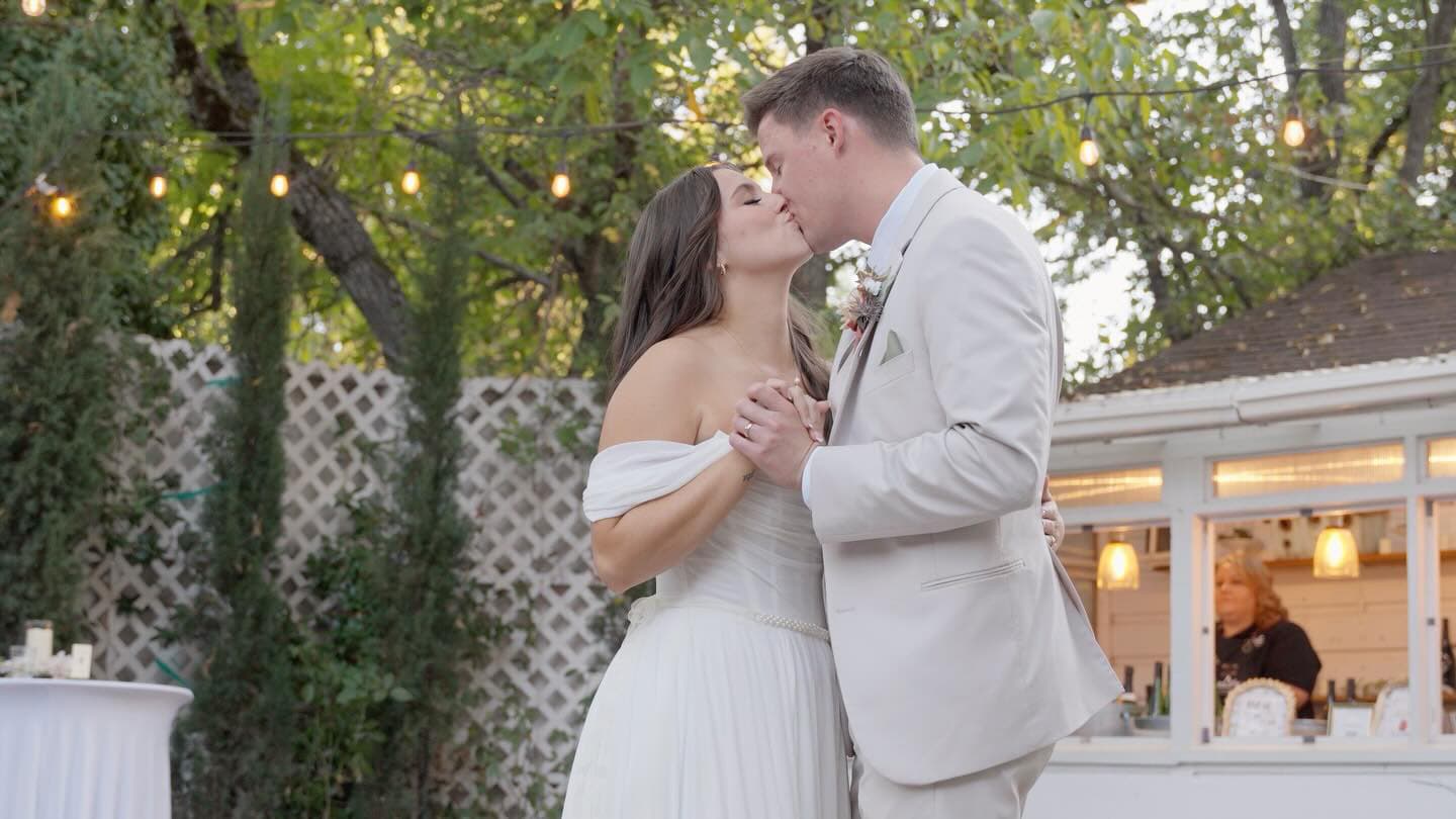 A couple shares a kiss during their wedding ceremony in an outdoor setting adorned with string lights.