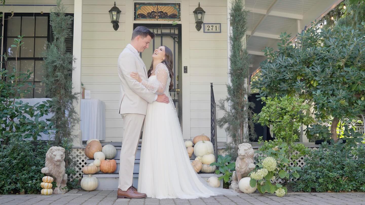 A couple embraces in front of a charming house adorned with pumpkins and greenery.