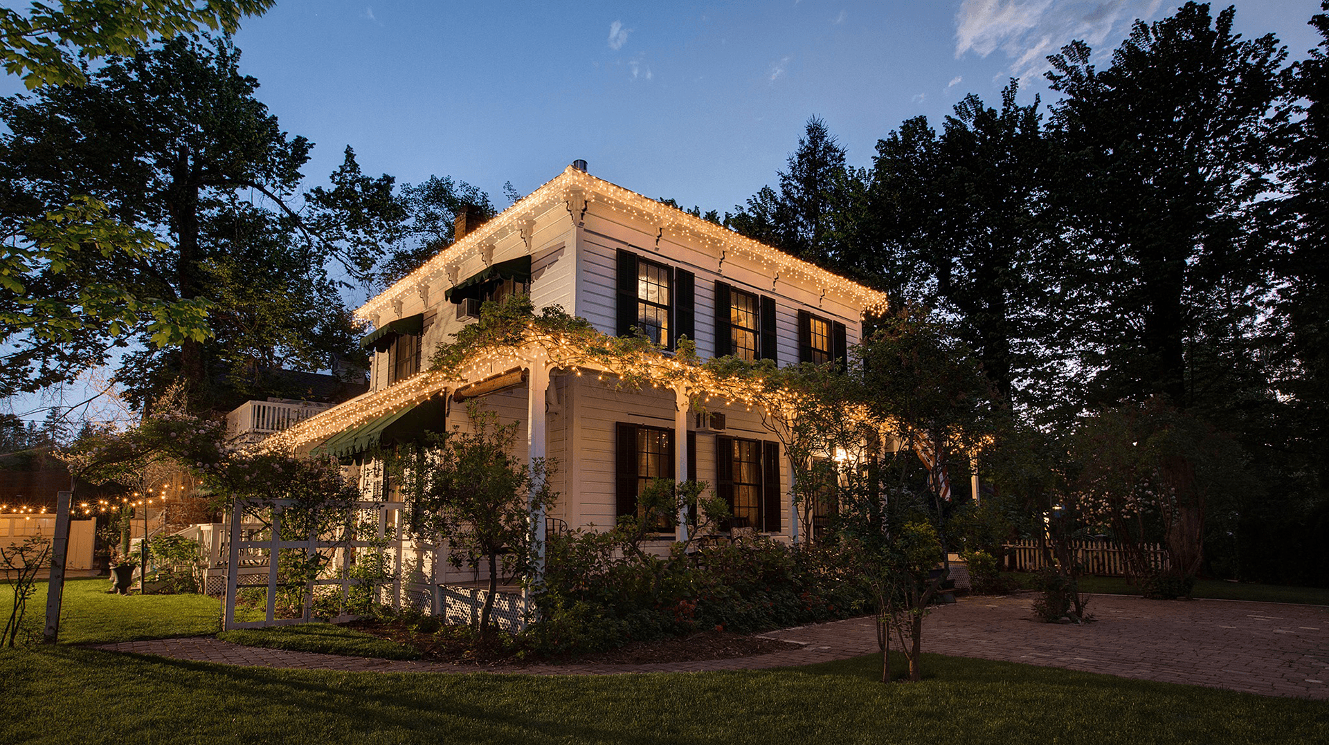 A charming two-story house adorned with warm lights, surrounded by greenery at dusk.
