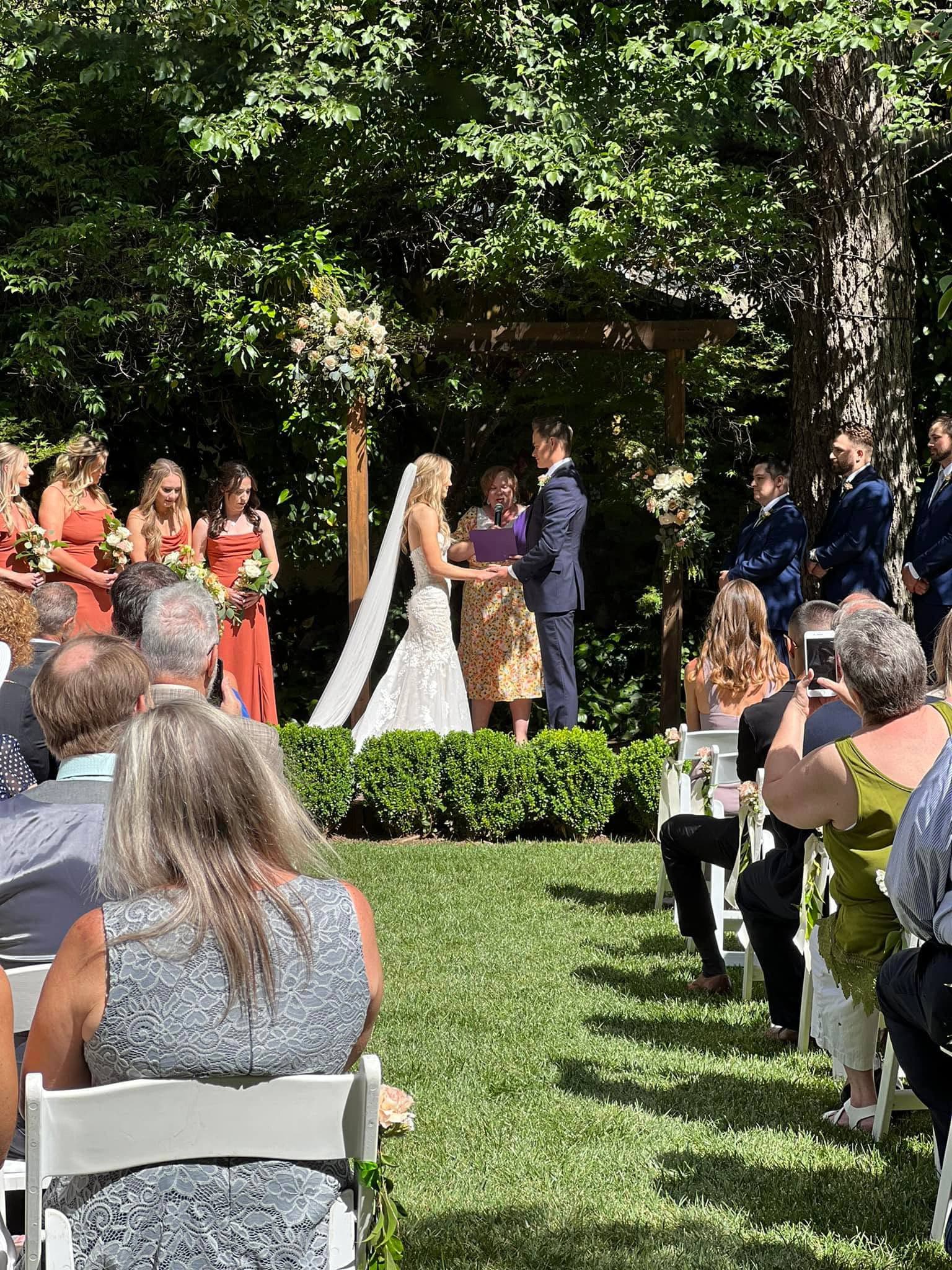 A bride and groom exchange vows under a floral arch during an outdoor wedding ceremony surrounded by guests.
