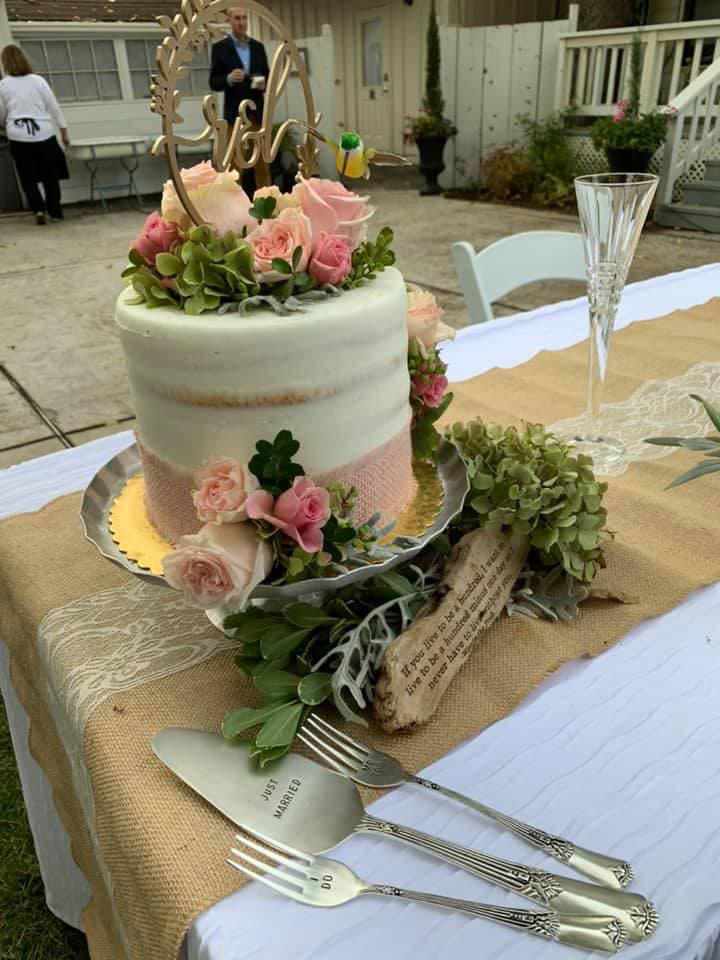 A beautifully decorated wedding cake topped with flowers and a "Just Married" sign, accompanied by elegant cutlery on a rustic table.
