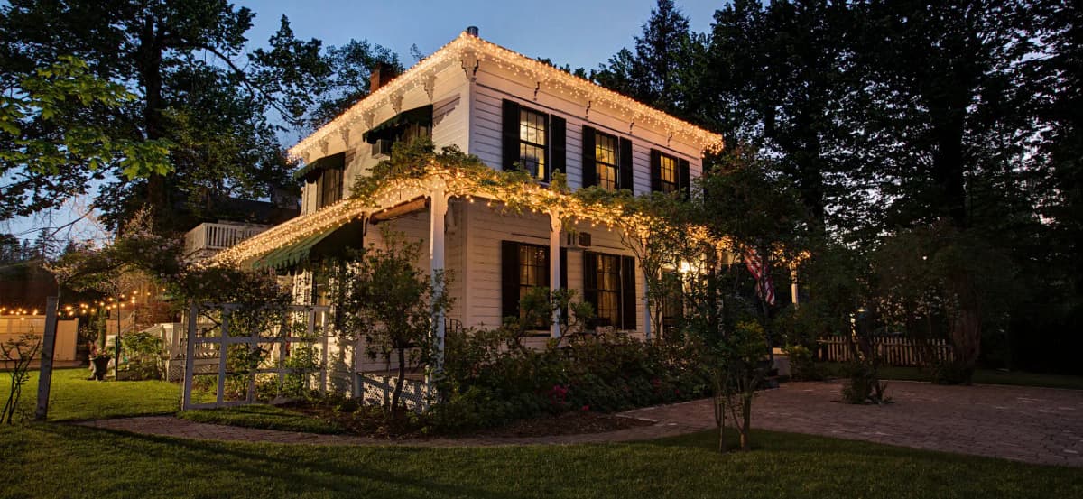 exterior of two story white building with black shutters, white string lights around edge of roof on two levels, green lawn in foreground