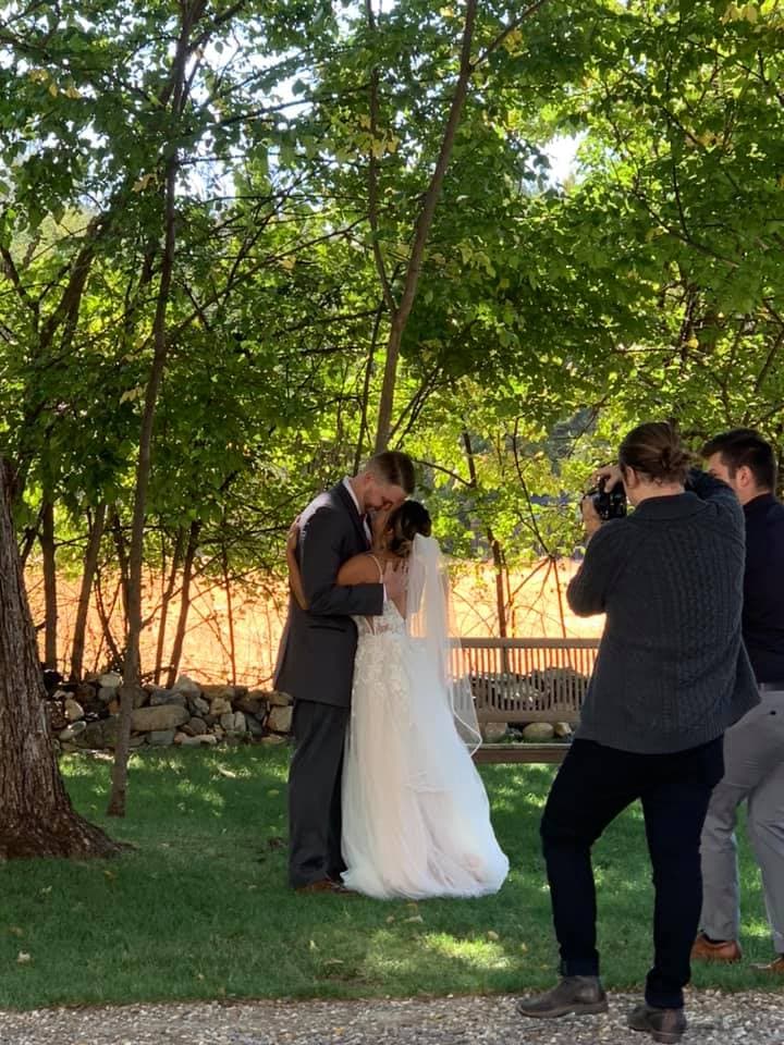A bride and groom kiss under a tree while being photographed by a videographer.
