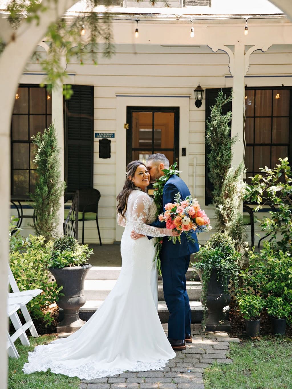 A bride and groom embrace joyfully in front of a charming house, surrounded by greenery and flowers.