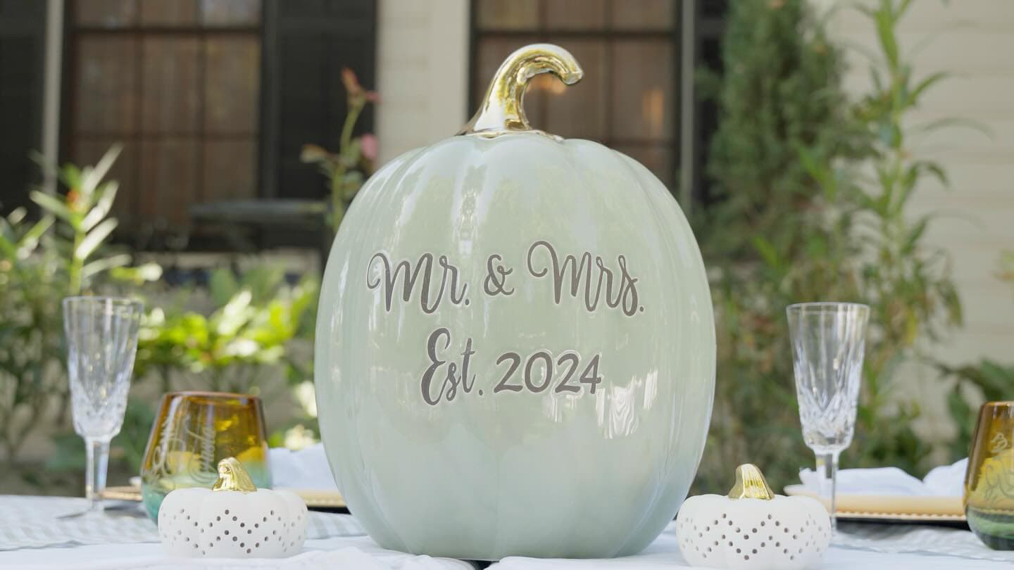 A decorative pumpkin inscribed with "Mr. & Mrs. Est. 2024" sits on a table surrounded by elegant glassware.