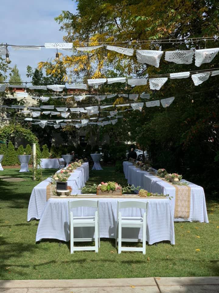 A beautifully arranged outdoor dining setup with white tablecloths, floral centerpieces, and decorative papel picado hanging overhead.