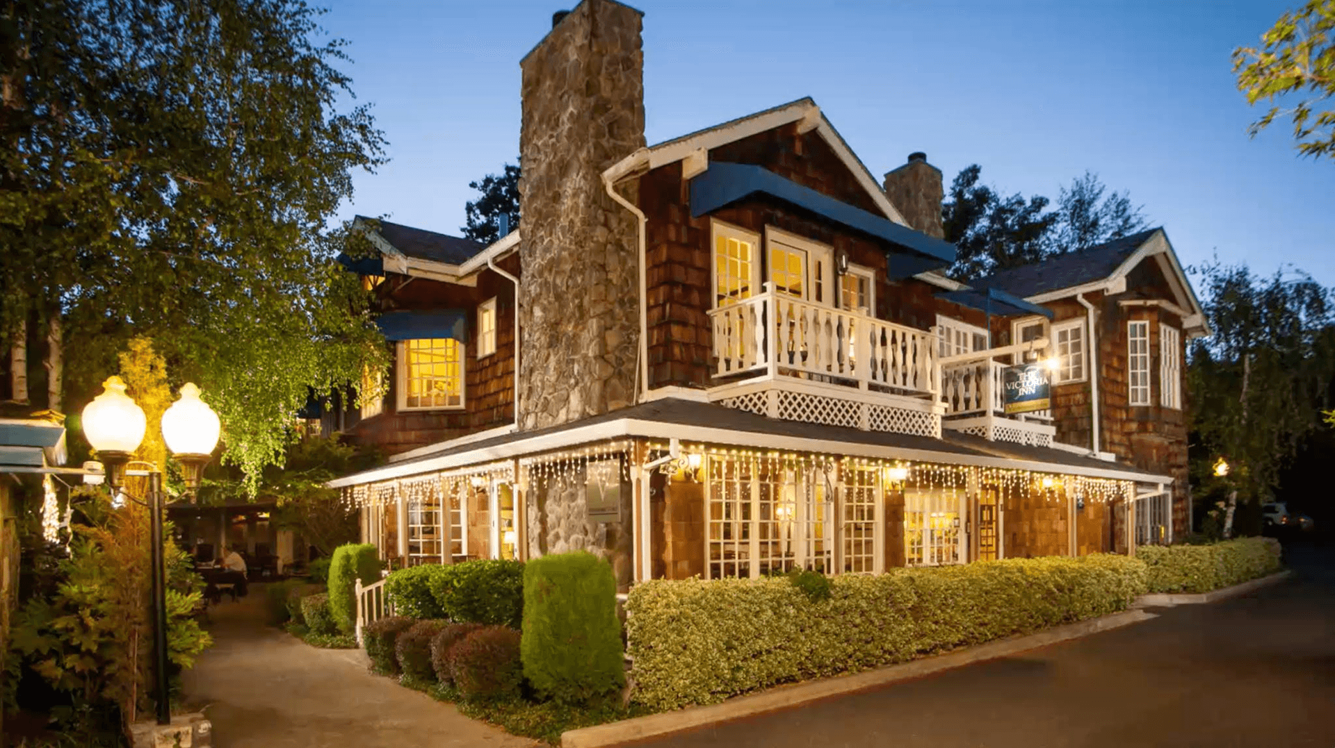 A charming two-story wooden house with a stone chimney, illuminated by soft lights and surrounded by lush greenery.