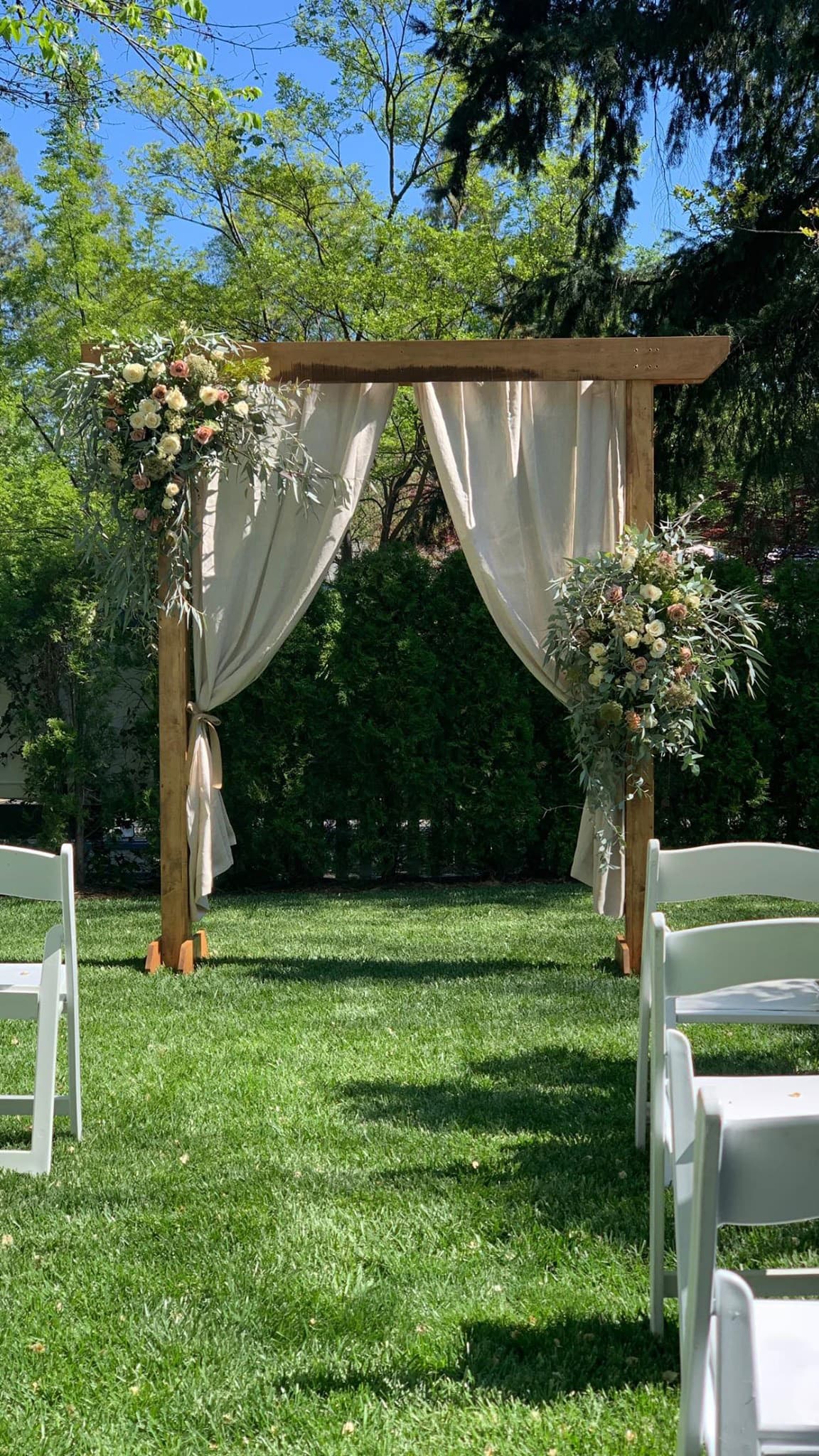 An elegant wedding arch with draped fabric and florals, set on a grassy area surrounded by lush greenery.