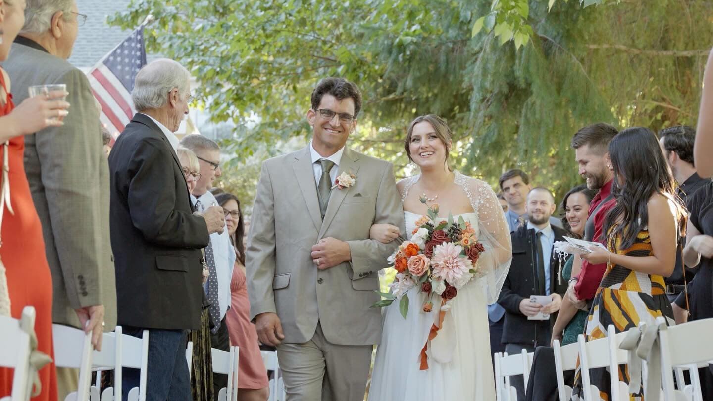 A smiling bride and her escort walk down an outdoor aisle adorned with flowers, surrounded by guests.