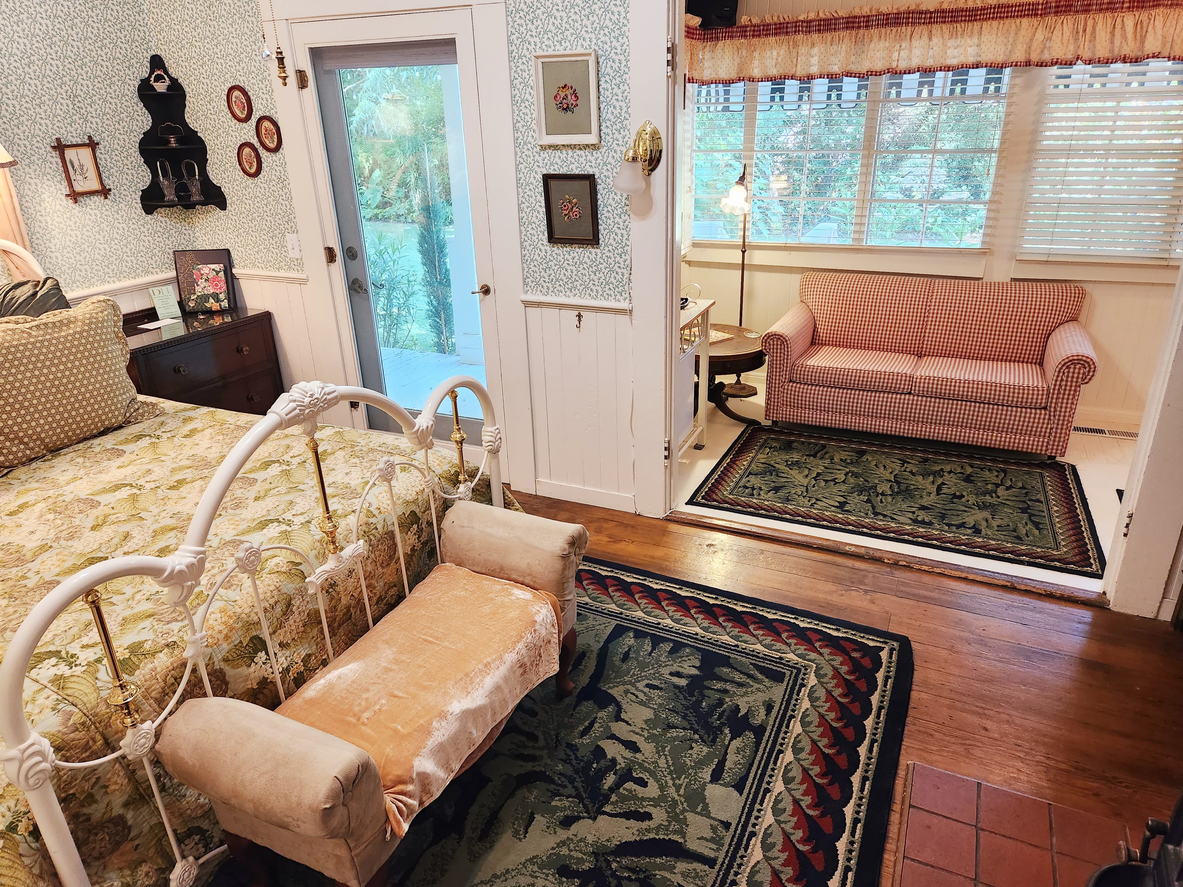 A cozy bedroom featuring a floral bedspread, vintage furnishings, a small sitting area, and large windows.