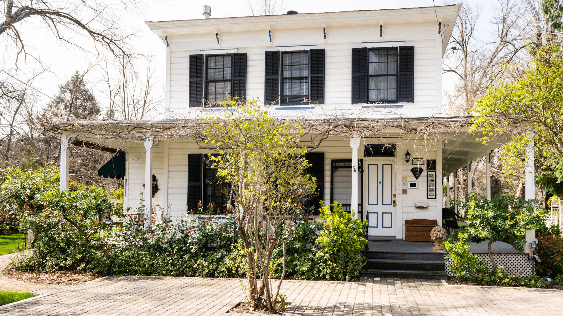 A charming white two-story house with black shutters surrounded by lush greenery and a brick pathway.