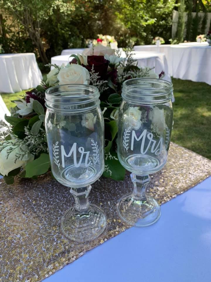 Two decorative glass goblets labeled "Mr." and "Mrs." sit beside a floral arrangement on a sequined tablecloth outdoors.