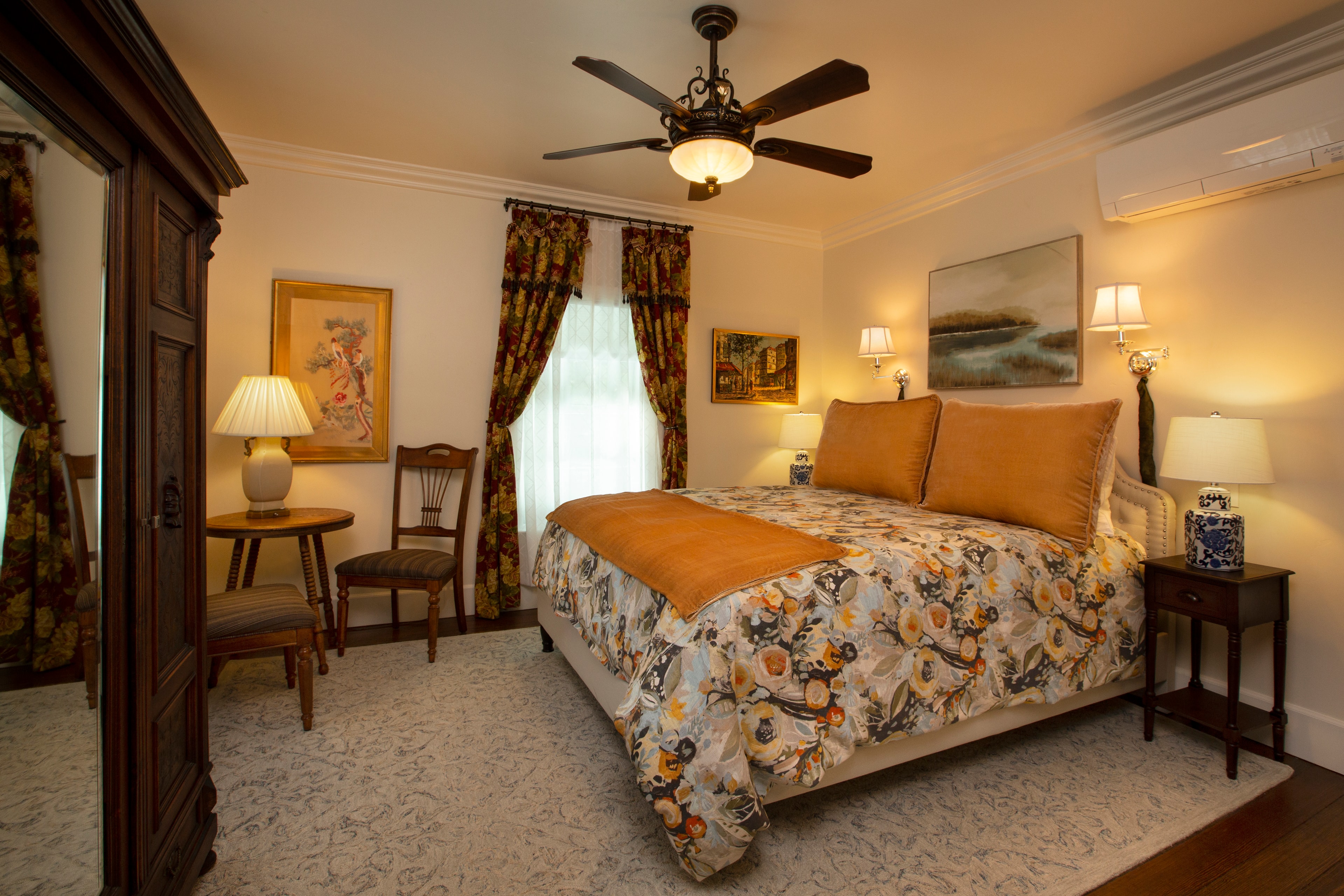 A cozy bedroom featuring a floral bedspread, two lamps, a wooden wardrobe, and two chairs by a window.