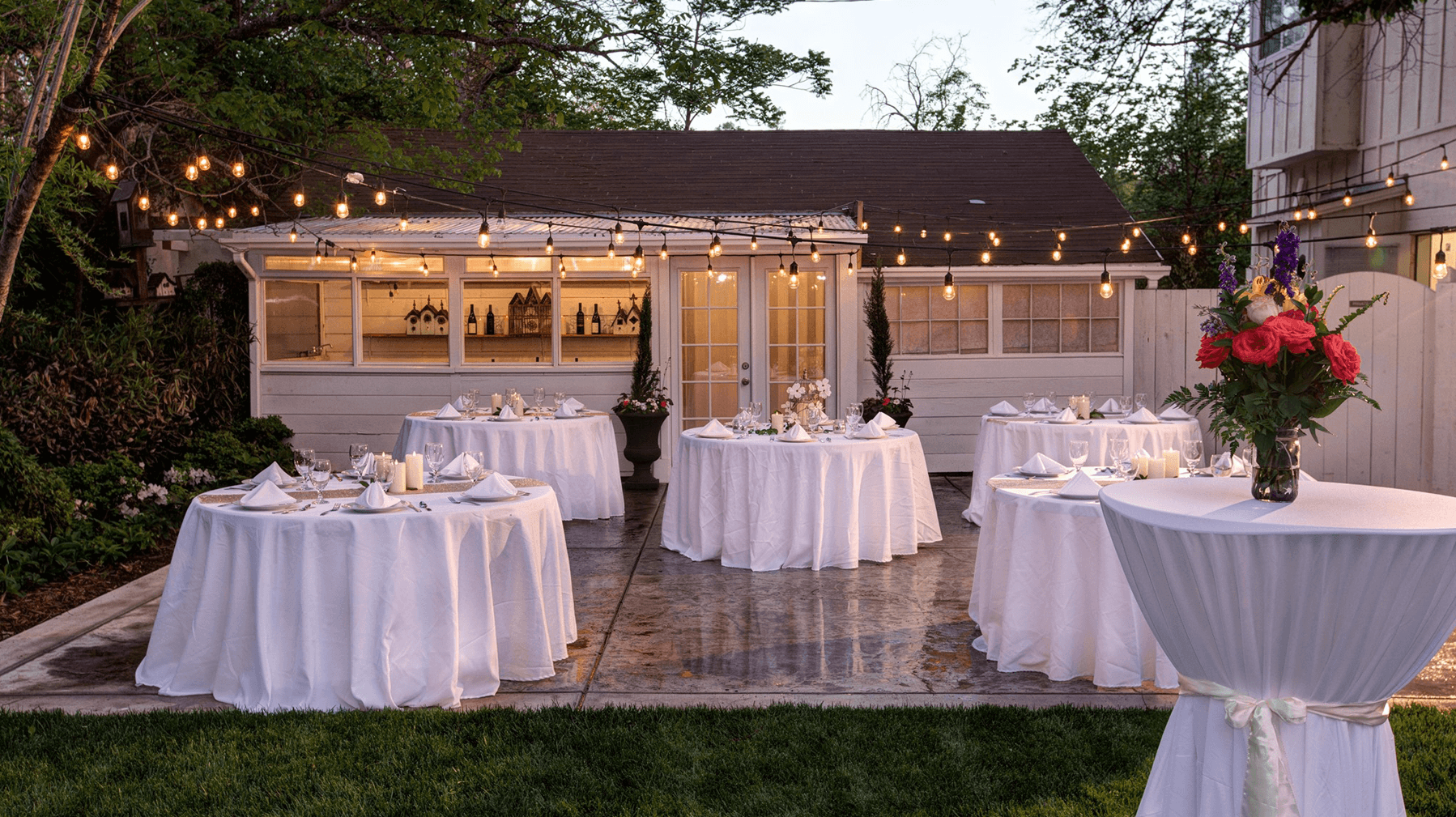 An elegantly set outdoor dining area with round tables, white tablecloths, and string lights, leading to a charming building in the background.