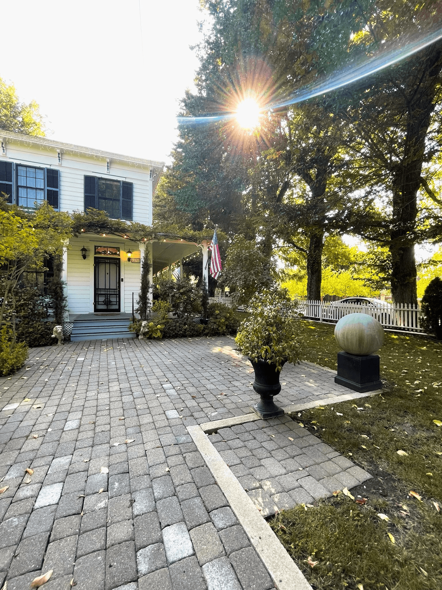 A charming white house with a front porch and American flag, surrounded by greenery and a sunburst through the trees.