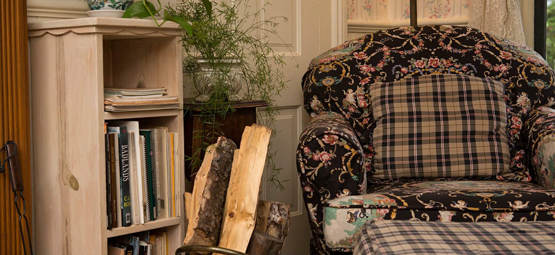 The corner of a living room with a small bookshelf with plants on it next to a large comfy sofa with plaid and floral patterns and firewood in between them. The corner of a living room with a small bookshelf with plants on it next to a large comfy sofa with plaid and floral patterns and firewood in between them.